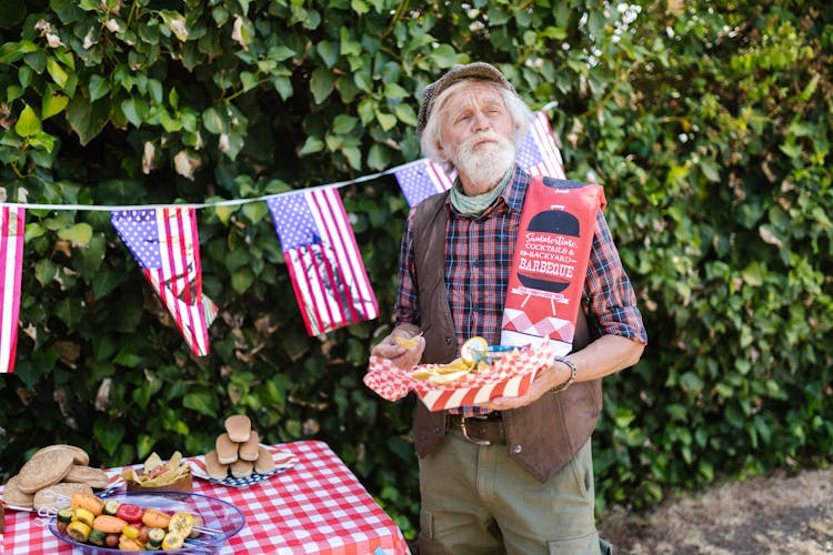 Elderly Man Holding A Tray With Food And Standing In The Garden Decorated For The 4th Of July