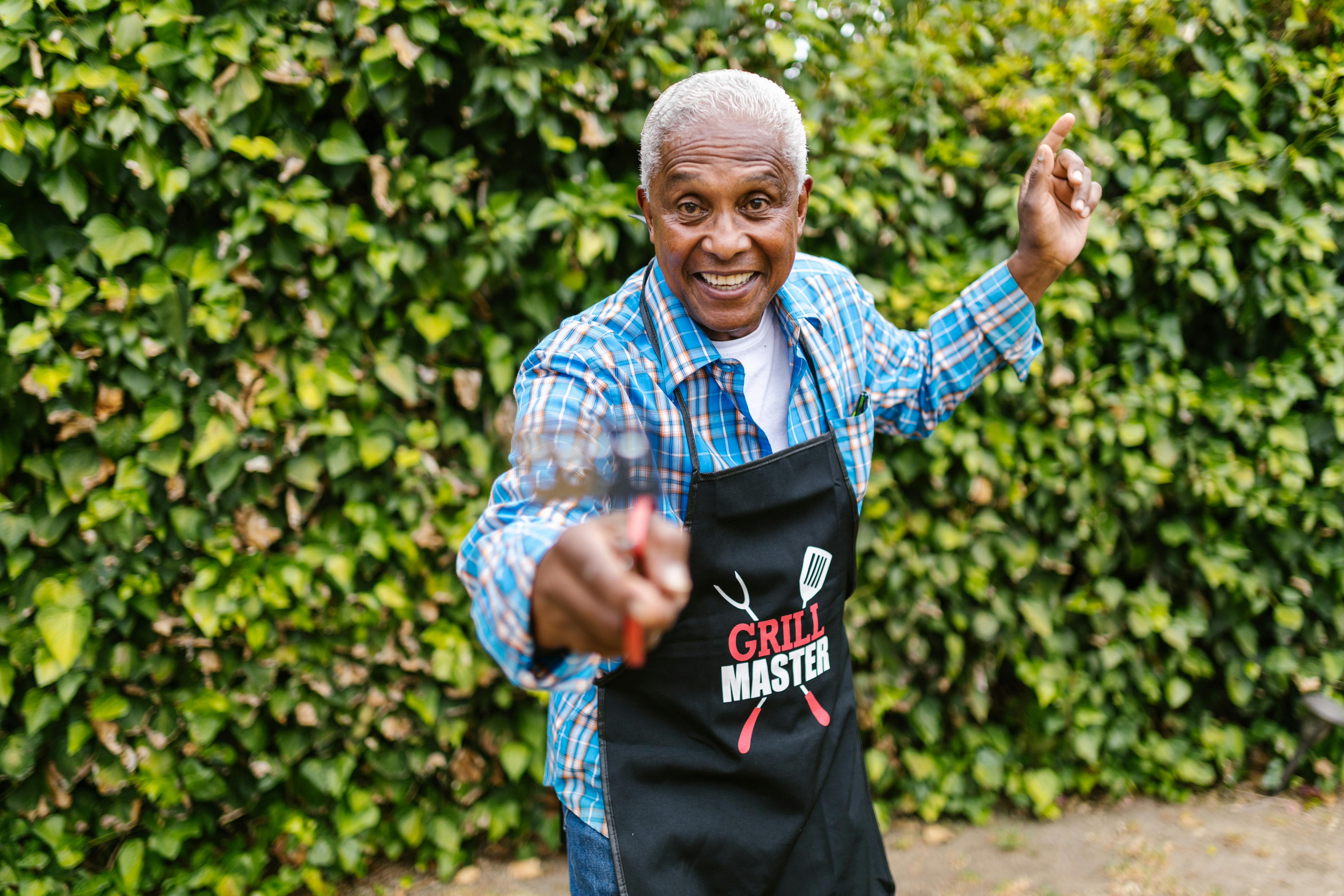 Cheerful elderly man in apron having fun while grilling in backyard with lush greenery.