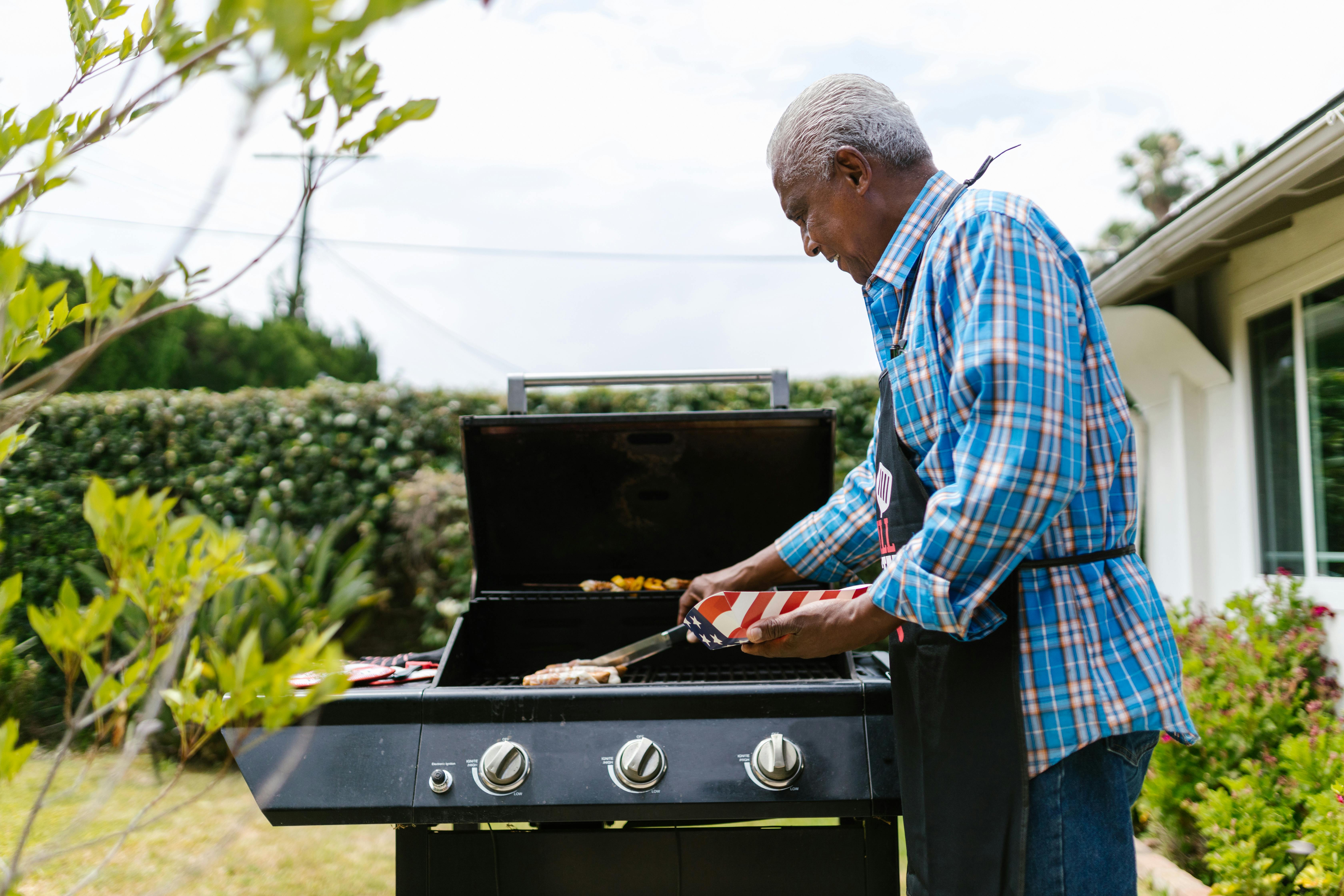 An elderly man grilling sausages on a barbecue in his backyard on a sunny day.