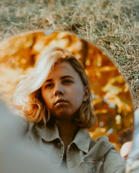 A young woman with blonde hair is seen in a mirror reflection outdoors, surrounded by autumn leaves.