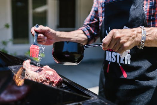 A person prepares a juicy steak on an outdoor grill, using a silicon brush to apply sauce.