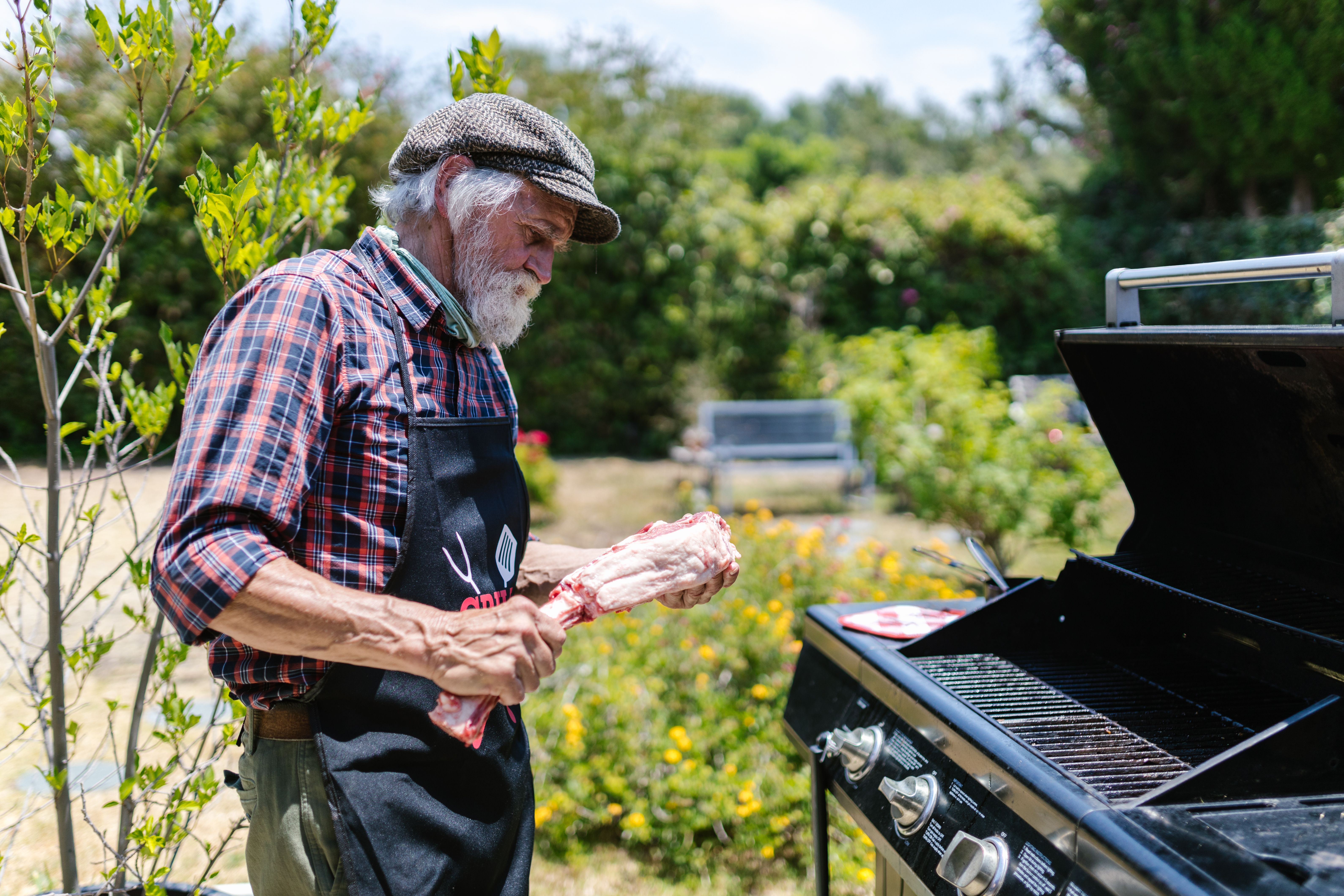 Senior man cooking meat on a gas grill in a sunny garden, enjoying a relaxed outdoor day.