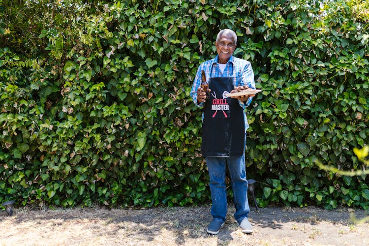 An Elderly Man In Plaid Long Sleeves And Black Apron Standing Near The Green Plants While Holding A Beer Bottle And A Plate With Food