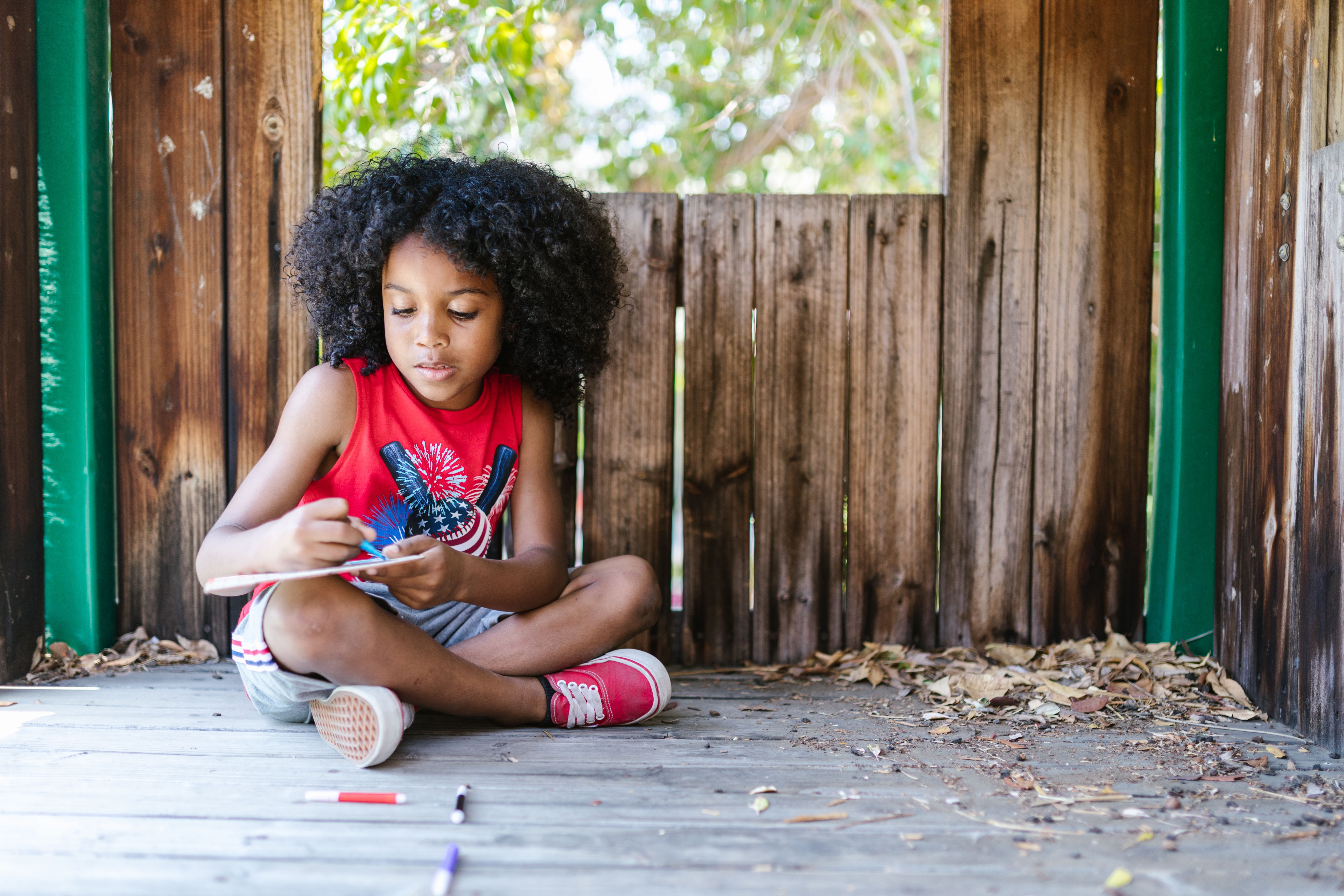 Young girl with curly afro hair drawing inside a wooden playhouse on a sunny day.