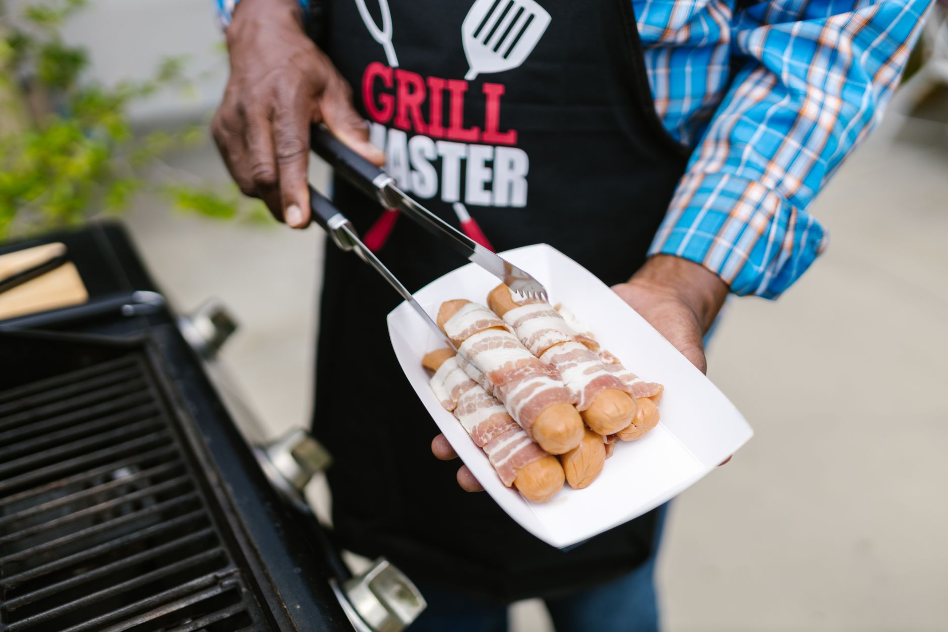 A person in an apron grilling bacon-wrapped hotdogs outdoors on a sunny day.