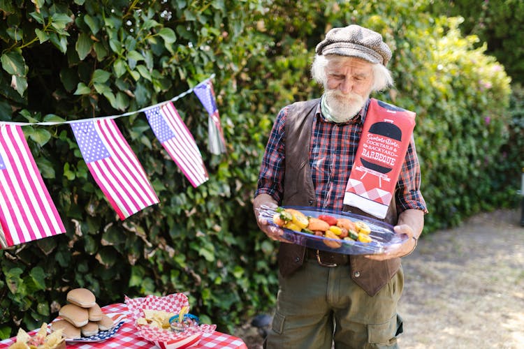 Elderly Man Holding A Tray With Food And Standing In The Garden Decorated For The 4th Of July