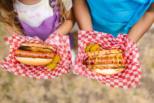 Close-up of kids holding grilled hotdogs with pickles and condiments outside.
