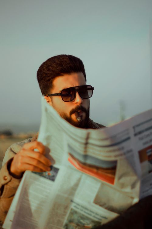 Free A fashionable man with beard and sunglasses reads a newspaper outside. Stock Photo