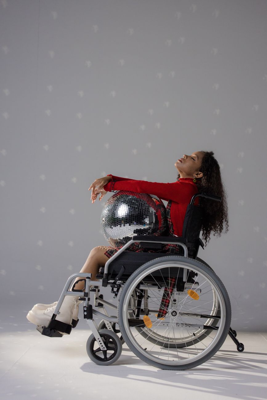 Jeune femme aux cheveux longs et bouclés, vêtue de rouge, assise dans un fauteuil roulant et tenant une boule à facettes, avec les yeux fermés et la tête penchée en arrière sur un fond gris.
