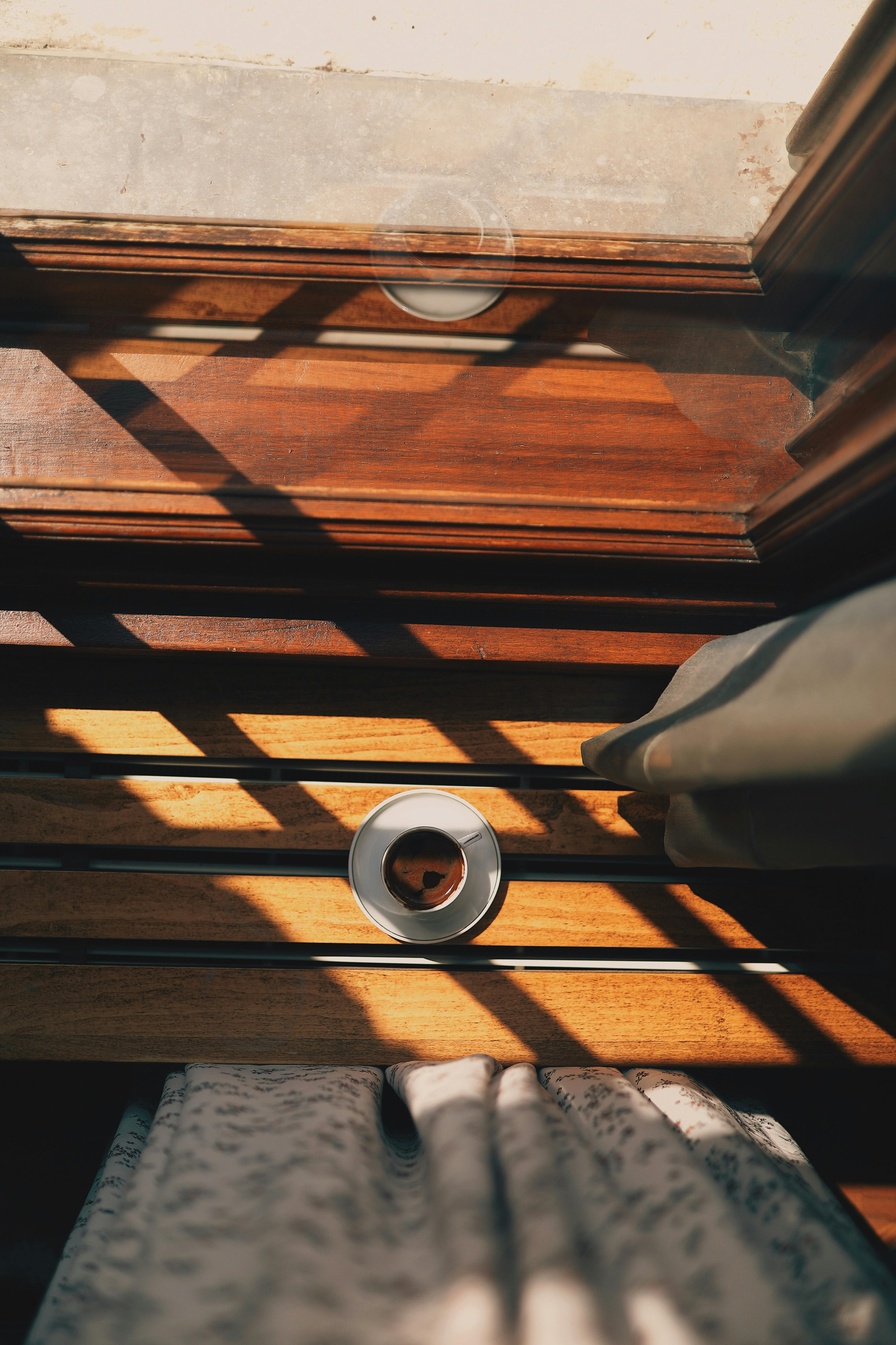 A warm cup of coffee on a sunlit wooden windowsill casting shadows.