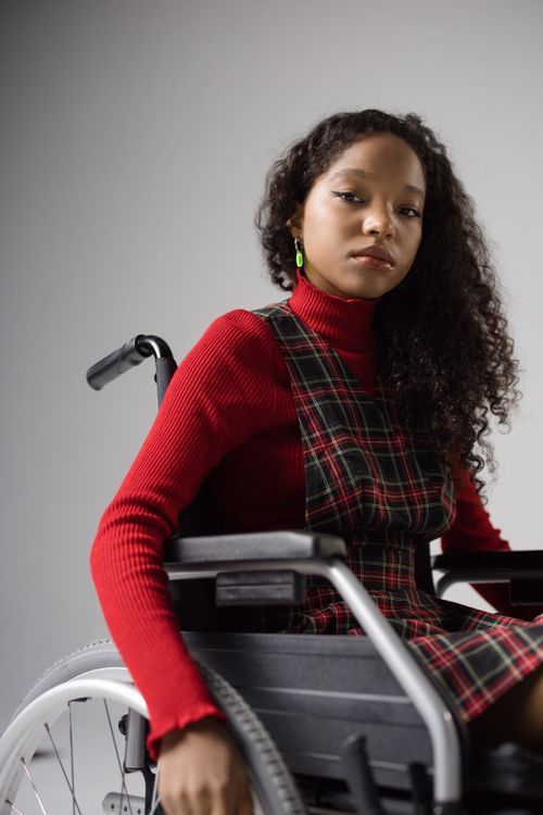 Free Portrait of a stylish young woman with curly hair in a wheelchair, wearing a red sweater and plaid dress. Stock Photo