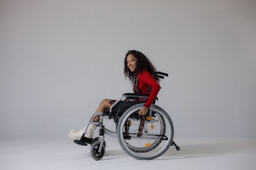 A young woman with curly hair smiling while seated in a wheelchair against a gray background.