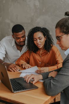 Three adults collaborating in an office setting, discussing documents with a laptop.