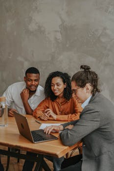 A diverse group of three adults working together around a laptop at a wooden table indoors.