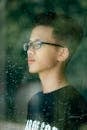 Through glass view of young pondering teen male in eyeglasses standing behind window with raindrops