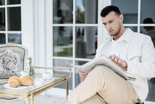Free Elegant man reading a newspaper while enjoying breakfast with croissants and coffee on a patio. Stock Photo