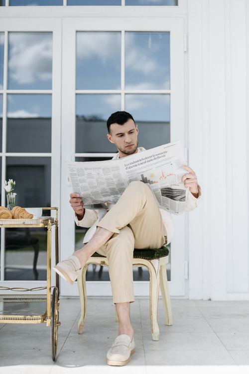 Free Stylish man in beige pants reading newspaper outside modern home. Stock Photo