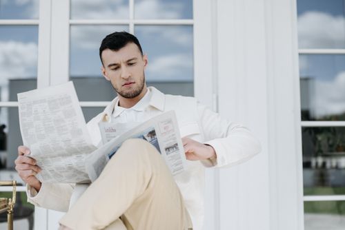 Free Young man in white attire reading a newspaper outside a building. Stock Photo
