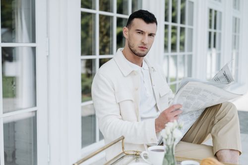 Free Stylish man in white and beige attire reading a newspaper outdoors. Stock Photo