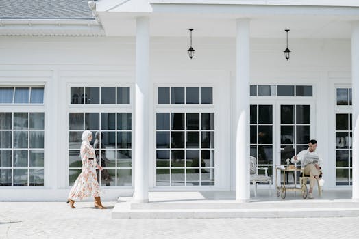 A man reads on a porch while a woman in a floral dress walks by. Modern exterior decor.