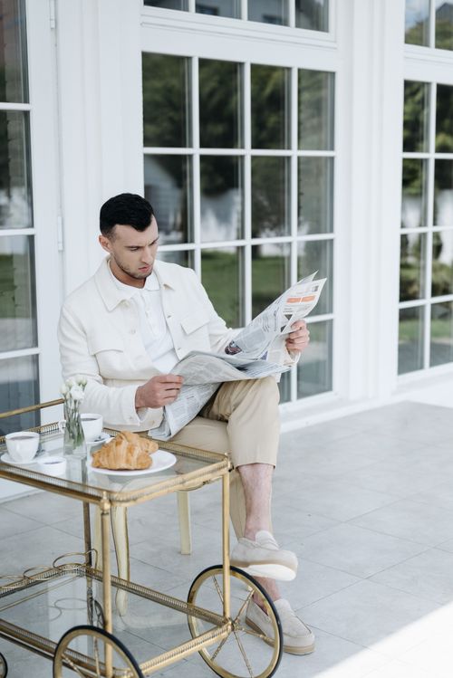 Free A man sitting in a sunroom, reading a newspaper by a chic brass table. Stock Photo