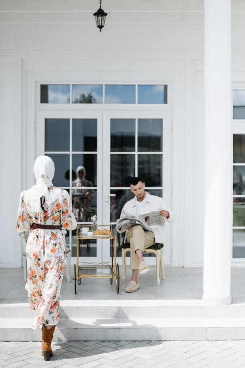 Woman in Floral Maxi Dress with White Headscarf Walking Towards a Man Reading Newspaper