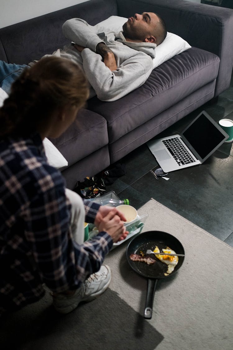 Sad Man Lying On A Sofa While Woman Getting Dirty Dishes