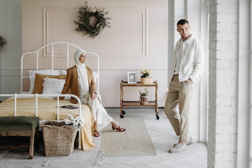 A Woman Sitting on the Bed while Looking at the Man Standing Near the Wall