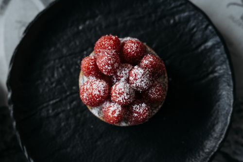 Free A vibrant raspberry tart with powdered sugar on a textured black plate. Stock Photo