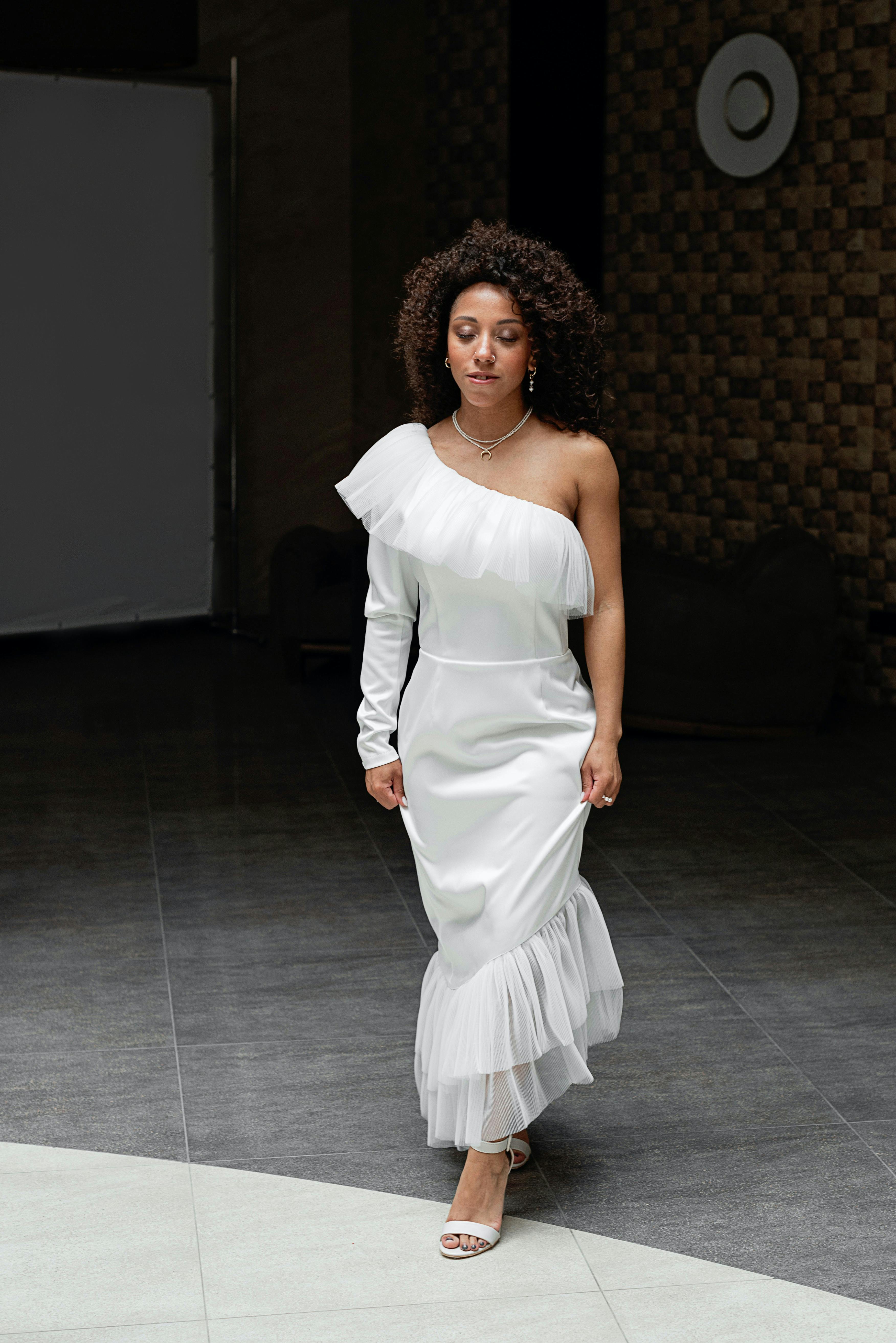 Beautiful African American bride walking indoors in a stylish off-shoulder white dress with curly hair.