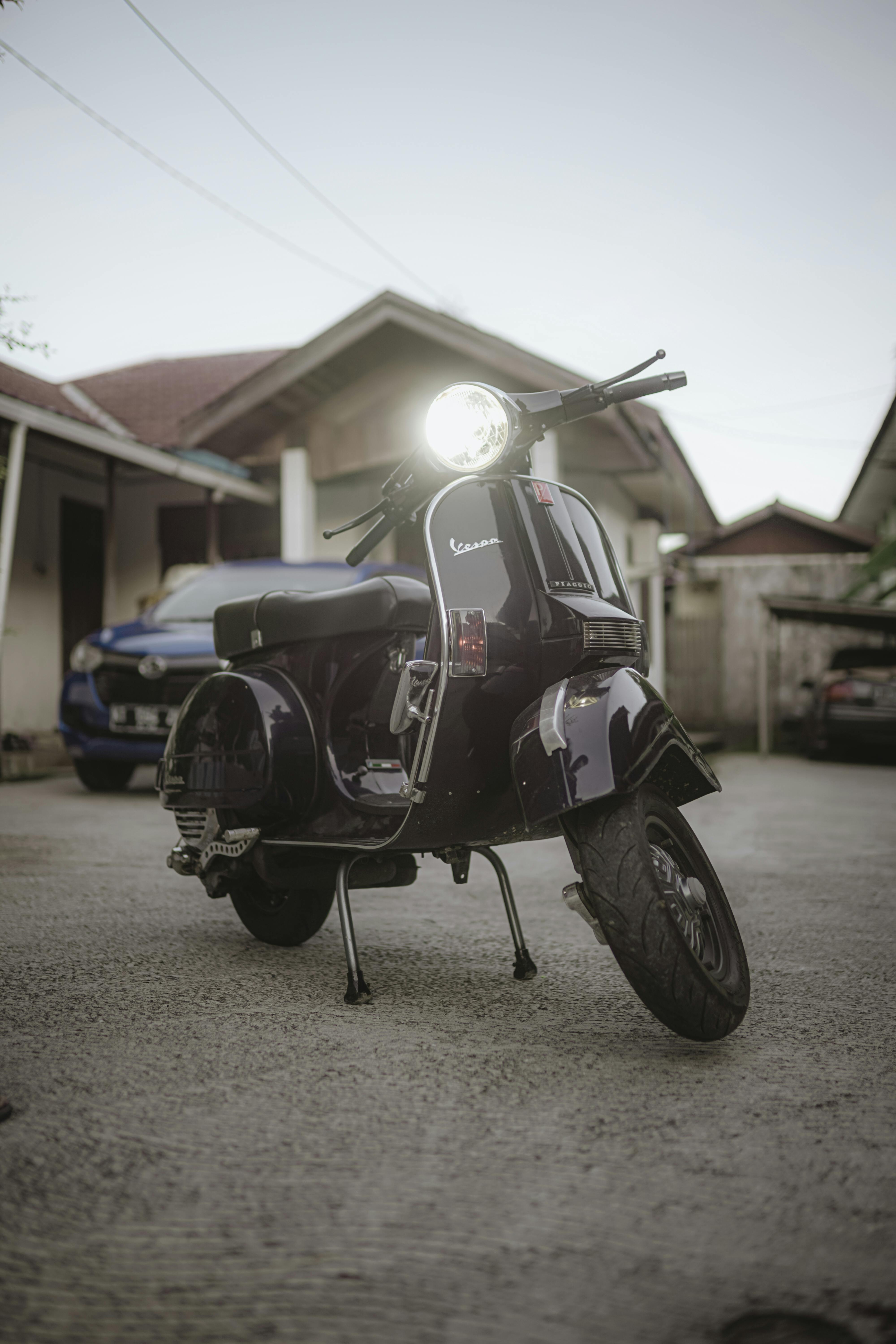 Classic black Vespa scooter with headlight on, parked outdoors against residential backdrop.