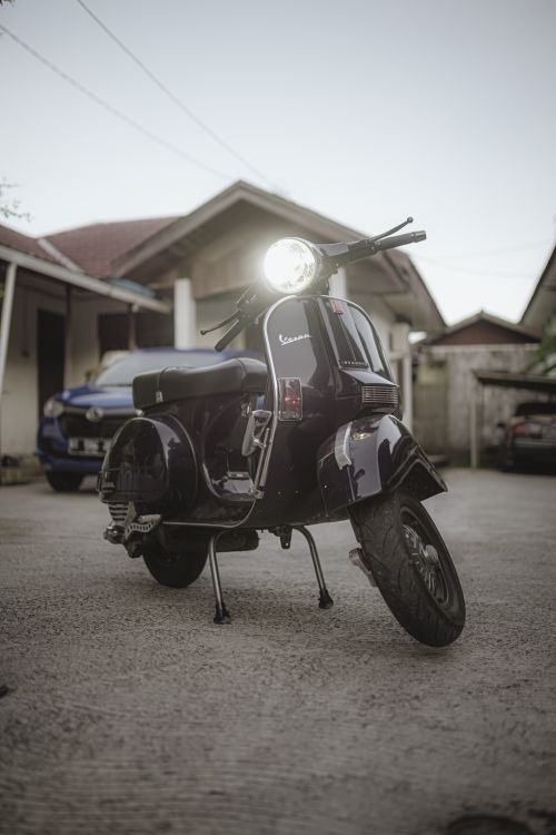 Free Classic black Vespa scooter with headlight on, parked outdoors against residential backdrop. Stock Photo