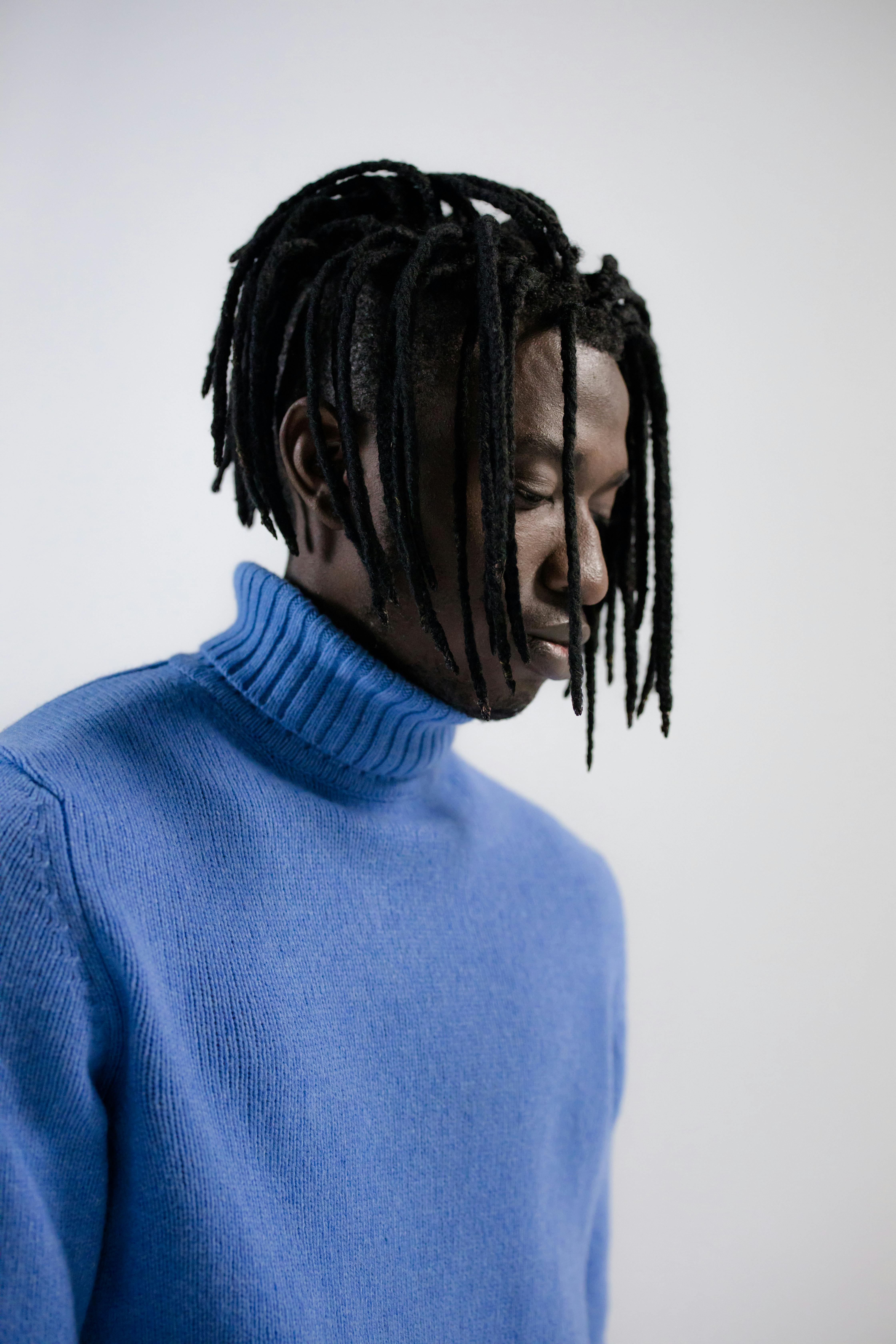 Portrait of a man with dreadlocks wearing a blue turtleneck sweater, looking thoughtful.