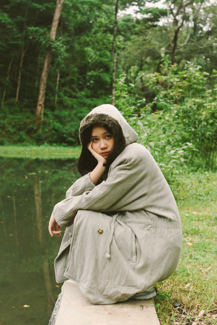 A woman in a green coat poses thoughtfully by a serene pond surrounded by lush greenery.