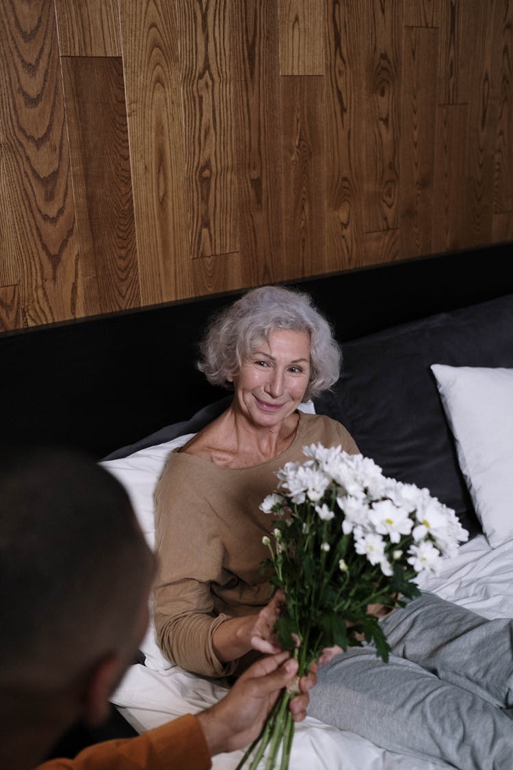 An Elderly Woman Sitting On The Bed While Holding Flowers