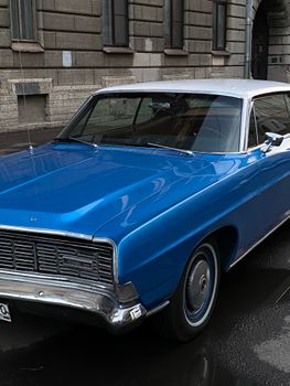 A classic blue vintage car parked on a rainy urban street in front of historic buildings.