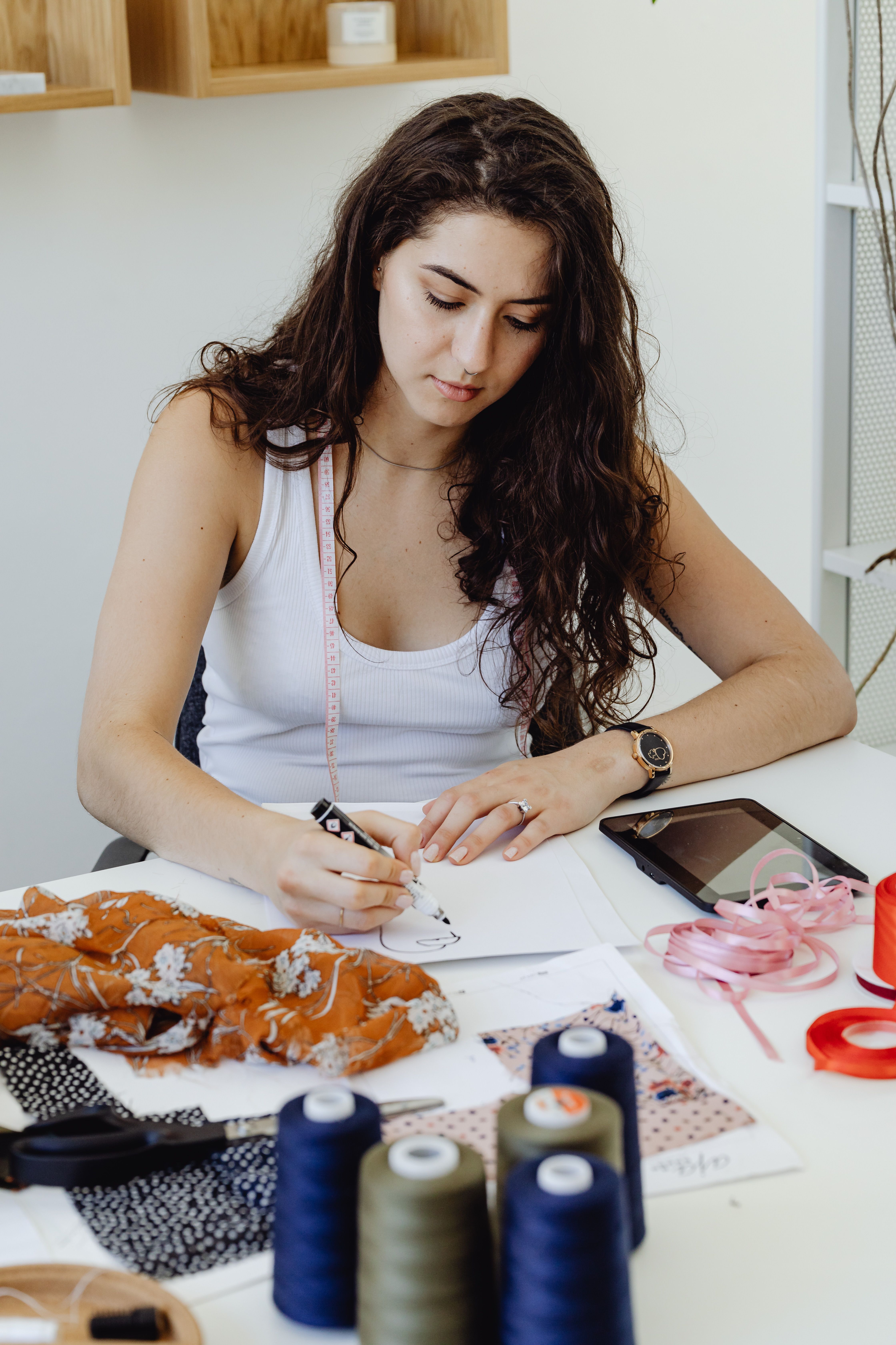 A young woman drawing fashion designs on paper at her workspace with fabrics and sewing tools.
