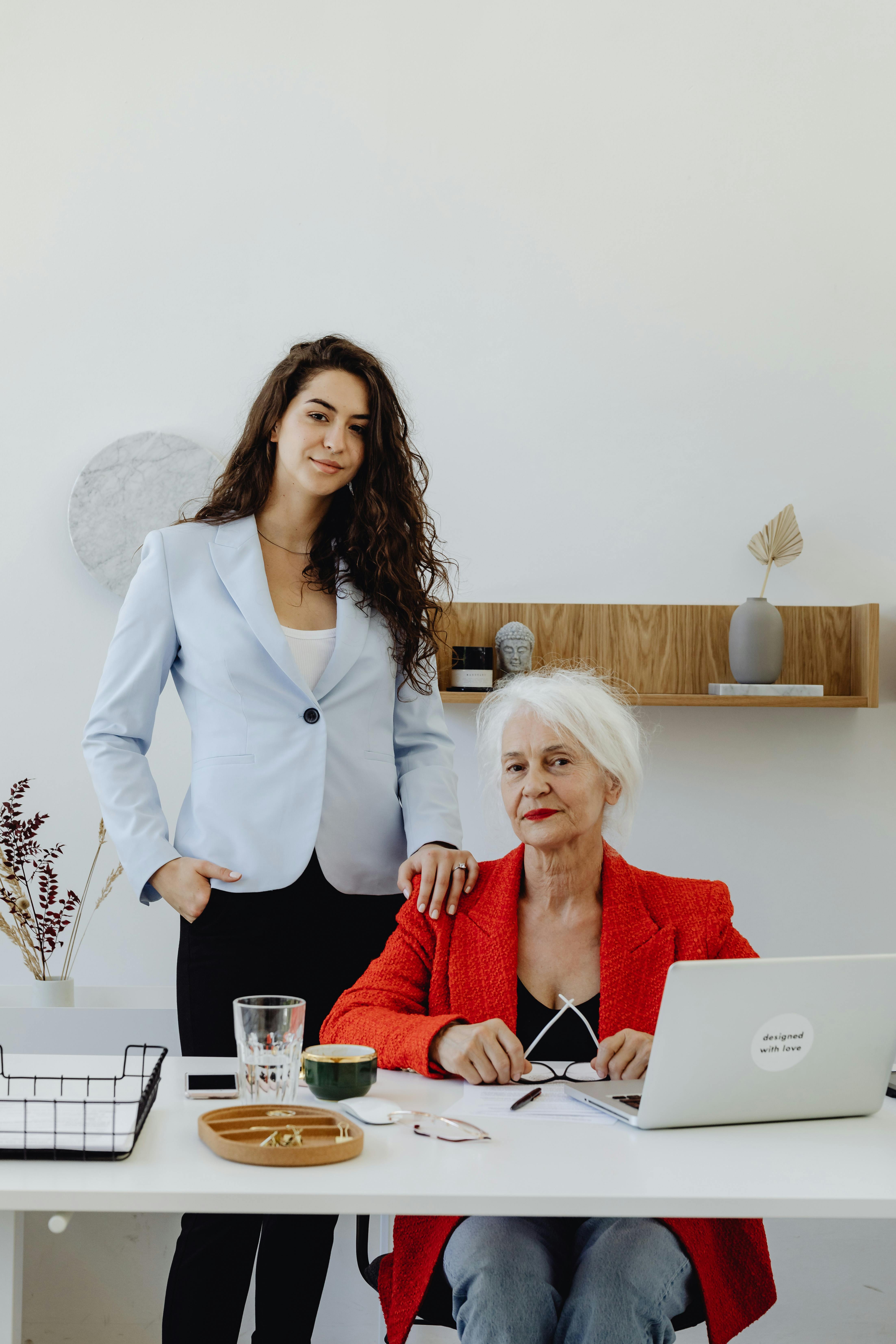 a woman in white blazer standing near the elderly woman in red blazer sitting near the table