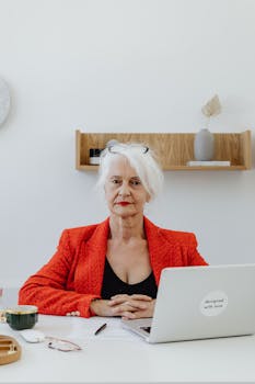 Senior businesswoman in red blazer confidently sitting at desk with laptop and cup, portraying professionalism.