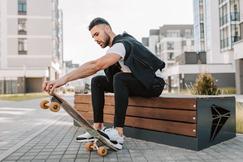 A Man in Black Pants Sitting on a Wooden Bench while Stepping on His Skateboard