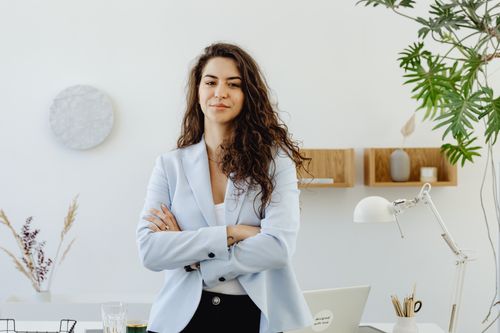 Une jeune femme aux cheveux longs et ondulés, vêtue d'un blazer bleu clair, pose les bras croisés dans un bureau lumineux et minimaliste agrémenté de plantes et d'objets de décoration.