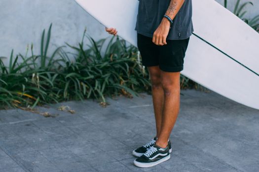 A man standing casually on a sidewalk holding a surfboard, embodying relaxed surf culture.