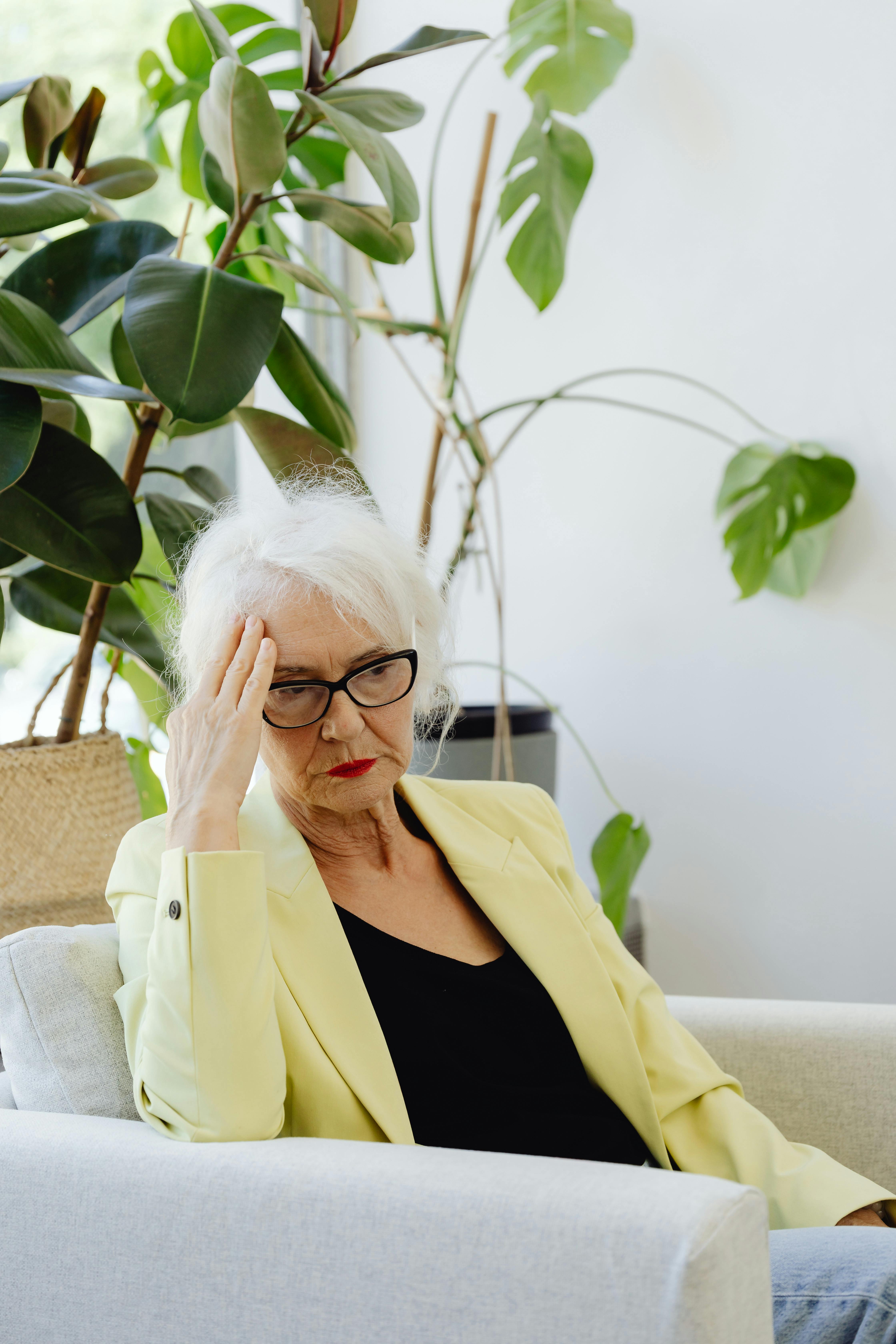 Elderly woman in yellow blazer, sitting and looking stressed with head in hand indoors.