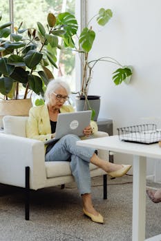 Elderly woman with white hair working on laptop in a plant-filled office space.