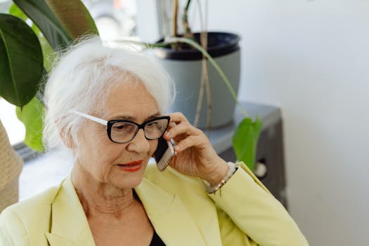 Elderly woman with white hair and eyeglasses using cellphone indoors.