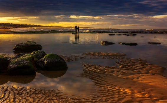 Couple walking on rocky shore at sunset with dramatic skies.