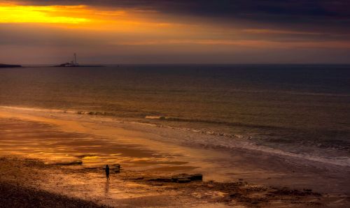 Free Peaceful beach at sunset with a silhouette of a person and lighthouse in the distance. Stock Photo