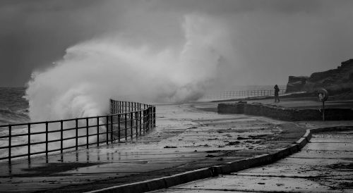 Free Powerful waves crash against a coastal path, fenced with metal railings, as a lone person walks amidst the stormy weather. Stock Photo