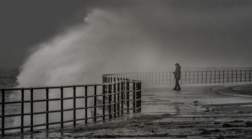 Free A lone man stands by railings as waves crash at a stormy seaside. Stock Photo