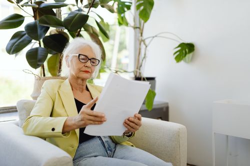 Free Elderly woman in yellow blazer reading papers at home near plants. Stock Photo